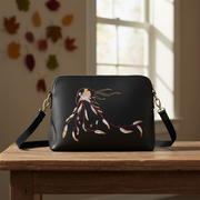 A black shoulder bag with a gold-tone strap and hardware, featuring a painted illustration of a woman with flowing hair and feather-like elements, displayed on a wooden table in a softly lit room.