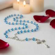 Glass Birthstone Rosary for March on a white surface with red petals and candles in the background.
