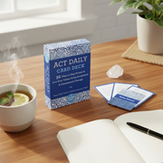 The 'Act Daily Card Deck' box is displayed on a wooden desk alongside a cup of tea with a lemon slice, an open notebook, a pen, and a small crystal, with a potted plant in the background.