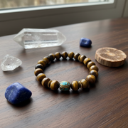 A beaded bracelet made of brown tiger's eye beads with a central turquoise and gold accent bead, displayed on a dark wooden table alongside raw crystals and a wood slice.
