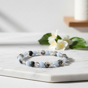 A beaded stretch bracelet with light blue, black, and clear faceted beads resting on a white marble tray, with white flowers and green leaves blurred in the background.