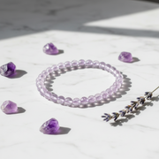 A translucent light purple beaded bracelet arranged in a circle on a white marble surface, surrounded by scattered purple crystal stones and a sprig of lavender under soft natural shadows.