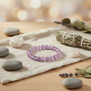 A purple beaded bracelet made of round beads, displayed on a beige textured cloth with spa props including smooth stones, a crystal cluster, a sage bundle, and dried leaves on a wooden surface.