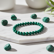 A green beaded bracelet made of polished round stones, displayed on a white marble slab with matching green stones scattered around it.