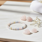 A pastel beaded bracelet made of round pearls in soft pink, blue, white, and pale yellow tones, arranged on a white marble surface with pink crystal stones and small dried flowers nearby.