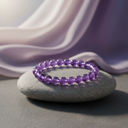 A purple beaded bracelet made of faceted oval beads, resting on a smooth gray stone with draped lavender fabric in the background.