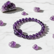 A purple amethyst beaded bracelet with a large polished oval bead, displayed on a white marble surface surrounded by scattered amethyst crystals.