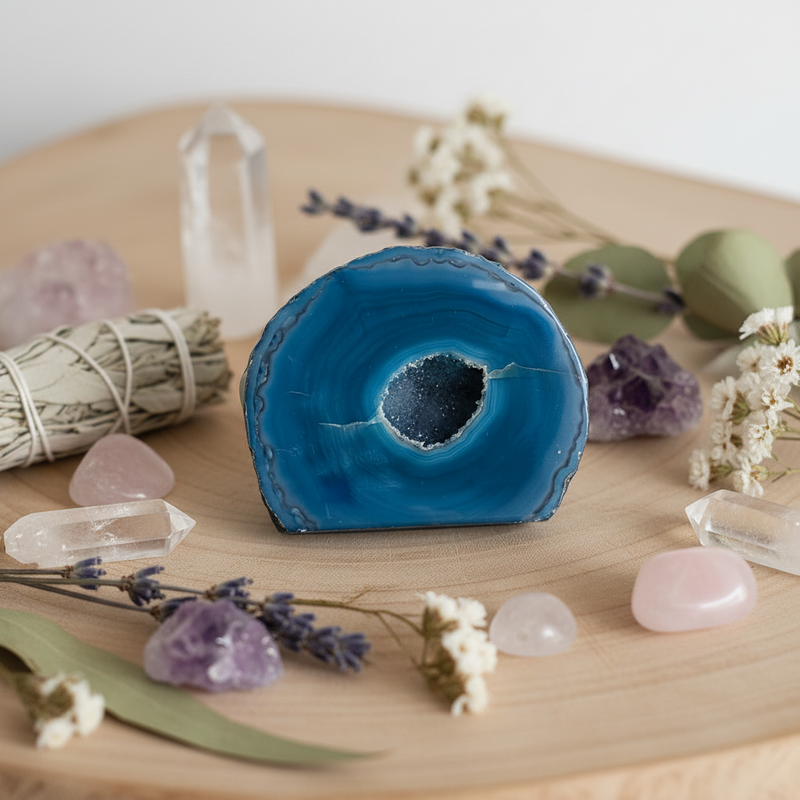 A polished  agate geode crystal with a hollow center, styled on a round wooden tray with quartz points, rose quartz, amethyst, lavender sprigs, dried flowers, and a bundle of sage.