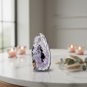 A polished purple dyed agate geode stands on a white marble tabletop in a softly lit room, with blurred pink candles and greenery in the background.
