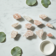 Several rough pale pink and clear crystal stones arranged on a white marble surface with green leaves and a small white cup in the corner.