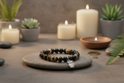 A beaded bracelet with dark brown and black tiger's eye beads and small metallic spacer rings, resting on a gray stone tray. The bracelet has a silver-toned charm.