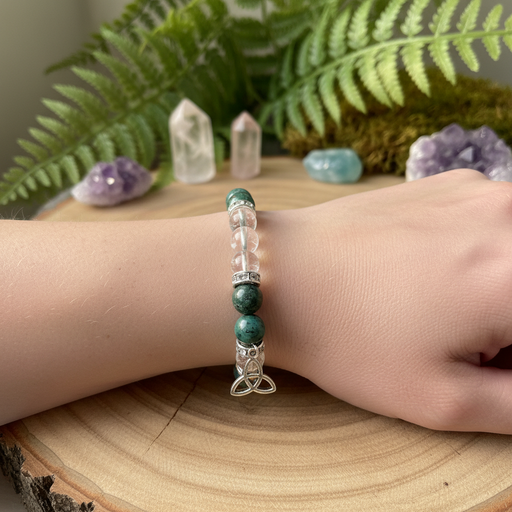 A beaded bracelet worn on a person's wrist, featuring green stone beads, clear crystal beads, and a silver Celtic knot charm, displayed on a wooden surface with crystals and ferns in the background.