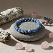 A blue beaded bracelet made of polished round stones sits on a smooth gray stone, surrounded by rose quartz crystals, a sage smudge bundle, dried lavender, and a second sage bundle on a beige linen surface.