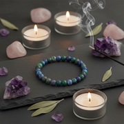 A beaded bracelet made of blue, green, and purple stones sits on a dark slate tray, surrounded by lit tealight candles, rose quartz stones, amethyst clusters, and a smoking incense stick.