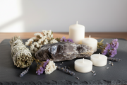 A styled arrangement of natural and spa-inspired items on a dark slate tray: a sage bundle tied with string, a smooth polished amethyst, dried white flowers, lavender sprigs, and three white pillar candles against a soft neutral background.
