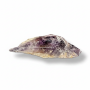 A raw, unpolished amethyst crystal, showing shades of purple, lavender, and clear quartz, displayed on a clean white background with a soft drop shadow.