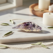 A amethyst crystal with purple and lavender tones, displayed on a white marble slab with dried leaves and candles in the background.