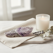 A lit white candle in a clear glass holder sits on a marble tray beside a purple amethyst crystal cluster, a beige linen cloth, and dried eucalyptus stems in a softly lit interior setting.