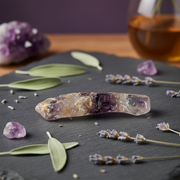 A polished amethyst crystal point tool lies on a dark slate surface, surrounded by lavender sprigs, green leaves, and small loose crystals. A blurred purple crystal cluster and a glass of amber liquid appear in the background.