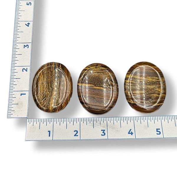 Three tiger's eye worry stones of varying sizes placed on a blue measuring tape, with the numbers 1 to 5 indicating the size scale.