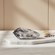 A rough polished amethyst crystal with natural purple, gray, and white marbling rests on a white marble tray, styled with dried grass and lavender against a beige paneled background.