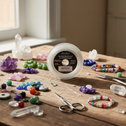A spool of clear craft cord or thread sits on a wooden table surrounded by colorful beads, crystals, small scissors, and other jewelry-making tools in a softly lit indoor setting.