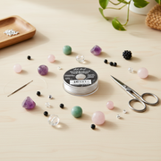 A spool of seed bead cord jewelry thread sits on a light wood table surrounded by loose beads, scissors, a needle, and small jewelry-making tools. The spool label reads 'Seed Bead Cord' and 'White'.