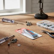 A tube of amazing goop adhesive in its retail packaging lies on a wooden workbench surrounded by screws, pliers, a screwdriver, and a small plastic tray of fasteners.