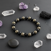 A beaded stretch bracelet made of alternating black and metallic gold beads, displayed on a dark slate surface surrounded by crystals and stones.