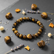 A beaded bracelet made of polished tiger's eye stones arranged in a circle on a dark slate surface, surrounded by matching loose beads, dried lavender sprigs, and small dried flower pieces.