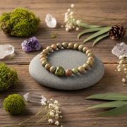 A beaded stretch bracelet made of marbled green, brown, and orange stones with a larger polished green stone, displayed on a smooth gray stone plate over a rustic wooden surface surrounded by moss, crystals, and dried flowers.