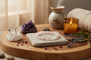 A pink and white beaded bracelet rests on a square stone slab on a round wooden tray, styled with crystals, a lit candle, a ceramic diffuser, and dried lavender sprigs.