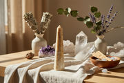 A tall, natural beige crystal tower stands on a draped cream linen cloth on a wooden table, surrounded by crystals, smudge bundles in ceramic vases, and a small wooden bowl with dried botanicals in warm natural light.
