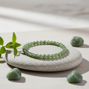 A green beaded bracelet displayed on a smooth light gray stone, with small green stones and a leafy sprig around it on a neutral beige surface.