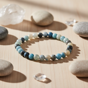 A beaded stretch bracelet made of round polished stones in shades of light blue, navy, gray, and cream, displayed on a light wood surface surrounded by smooth decorative stones and clear crystal points.
