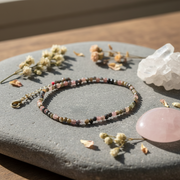 Multicolored beaded anklet on a stone surface with flowers and a crystal.