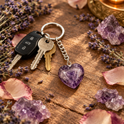A heart-shaped purple crystal keychain with a silver chain and split ring, attached to two keys, displayed on a rustic wooden surface surrounded by lavender, rose petals, and crystal clusters.