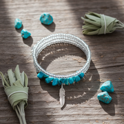 Bracelet with turquoise stones and feather charm on a wooden surface with turquois stones and sage.