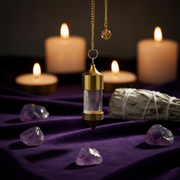 A gold and clear glass pendulum,  hangs from a chain on a draped purple fabric surface, surrounded by lit candles, purple crystals, and a white sage bundle in the background.